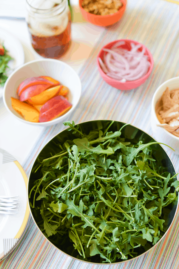 Bowl of arugula with smaller bowls with peaches and chicken next to it.