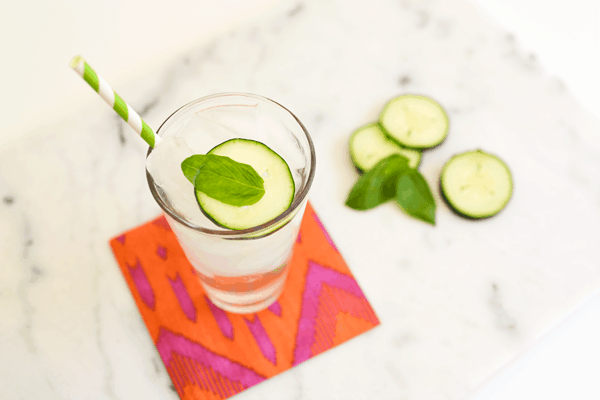 A glass with a mocktail sitting on an orange and pink napkin topped with  a cucumber slice and basil leaf.