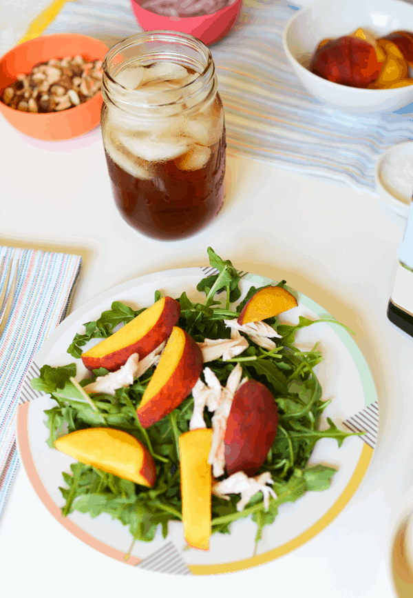 A salad with arugula, peaches, and chicken on a plate next to a Mason jar of iced tea. 