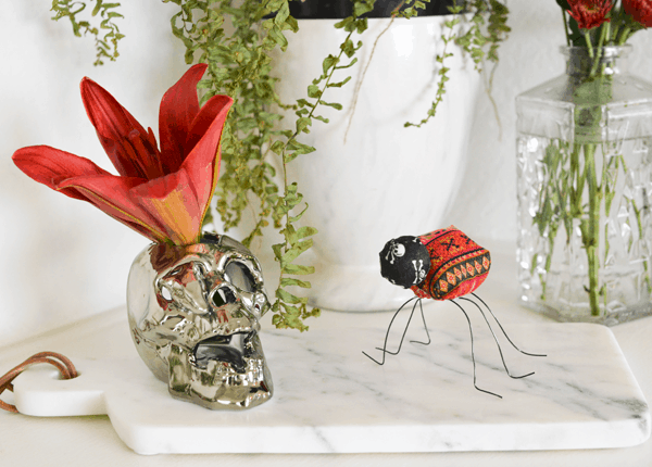 A homemade fabric bug with wire legs on a table next to a skull vase with an orange flower.