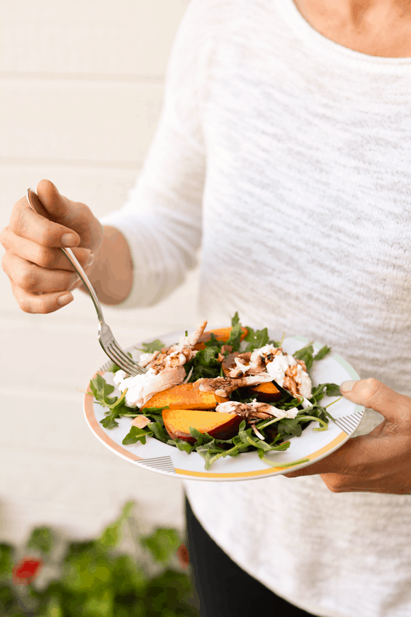 A woman holding a fork and a plate with an Arugula Salad topped with peaches and burrata and a balsamic drizzle. 