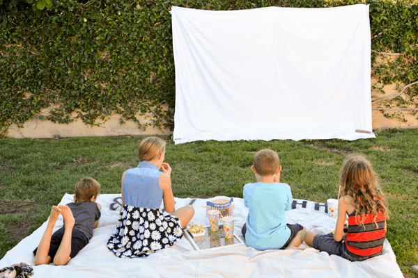 Kids sitting on the lawn in the backyard waiting for an outdoor movie to start on a sheet on the wall. 