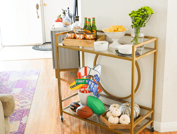 A bar cart at a home set up with snacks and drinks for a football tailgate party.