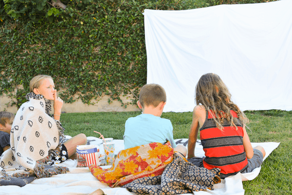 Kids sitting on the lawn in front of a. homemade movie screen. 