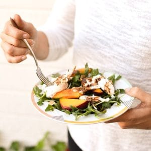 Woman eating a salad with peaches, burrata and balsamic on a bed of arugula.
