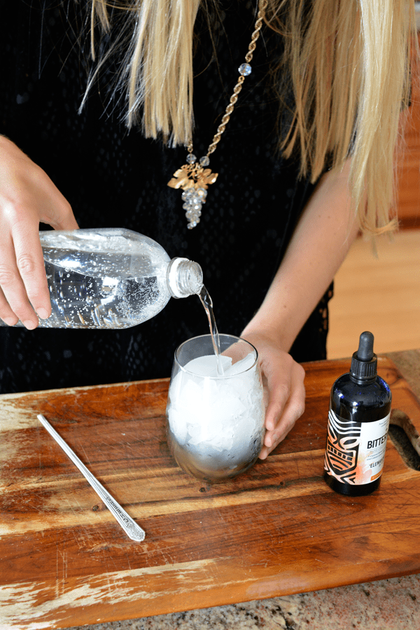 Woman pouring club soda into a stemless wine glass next to a bottle of bitters.