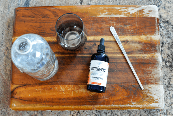 An overhead view of a cutting board with a bottle of bitters, a cocktail sitrrer and a stemless wine glass next to a bottle of club soda.