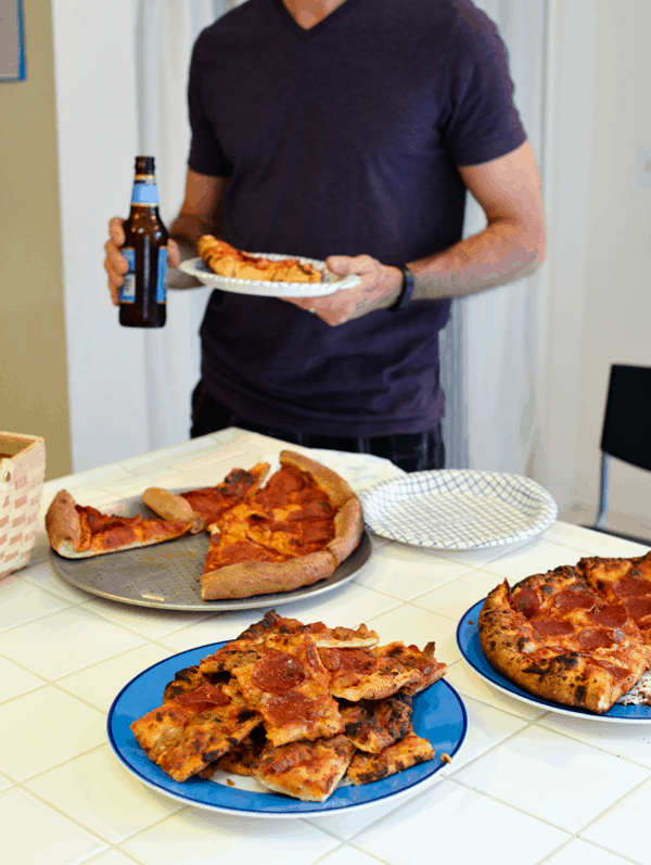 Pizzas on a counter with a man holding a plate of pizza and a beer in the background. 