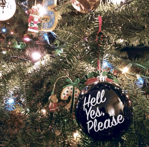 Close up of a black ornament on a Christmas tree that says, "Hell yes, please"