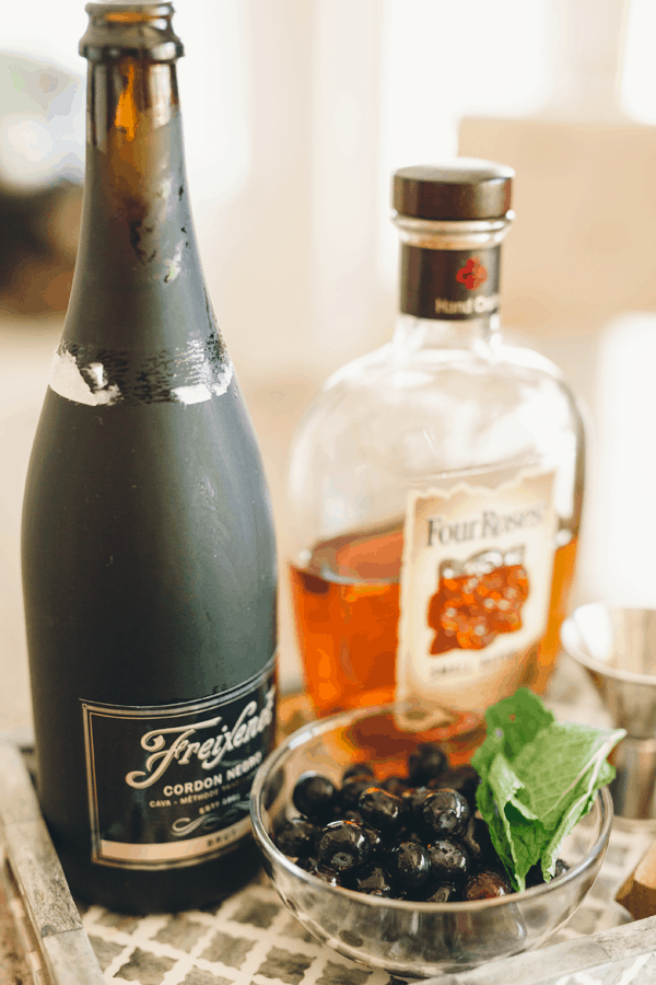A bottle of sparkling wine and bourbon on a tray with a small bowl of fresh blueberries and mint leaves. 