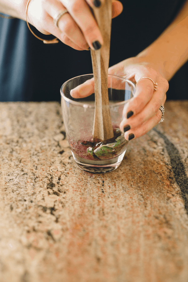 Woman muddling blueberries and mint in a short cocktail glass. 