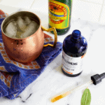 Bottle of bitters on a counter next to a copper Moscow Mule mug and a bottle of ginger beer.