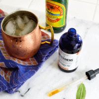 Bottle of bitters on a counter next to a copper Moscow Mule mug and a bottle of ginger beer.