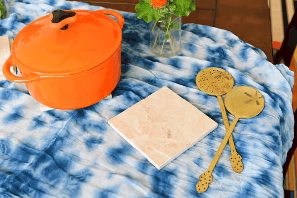 Table with orange covered baking dish next to a tile trivet and serving utensils.