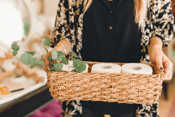 A woman holding a basket with toilet paper rolls and a few eucalyptus leaves.