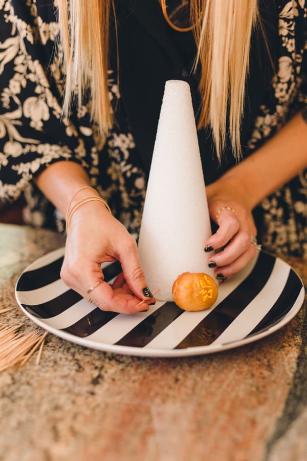 Woman adding a toothpick to a styrofoam tree form to make a donut tower.