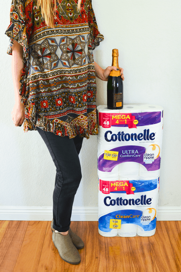 Woman standing next to two large packages of toilet paper resting a bottle of champagne on top.