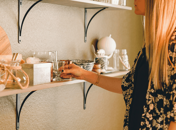 A woman standing in front of a wall shelf picking up a glass.
