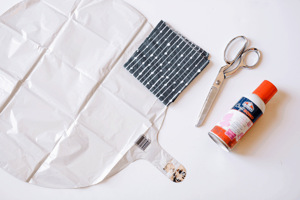 A white uninflated mylar balloon a black and white patterned napkin, spray adhesive and scissors laying on a table. 