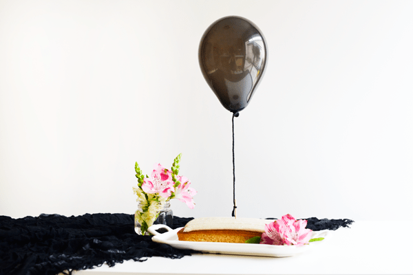 A small white platter with a 1/4 sheet cake with a toothpick holding yarn attached to a blown up black balloon.