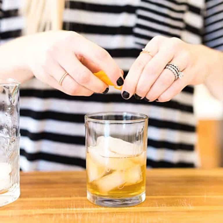 Woman adding an orange garnish to a Pine Old Fashioned cocktail on a counter.