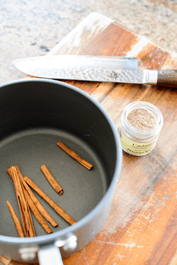Cinnamon sticks in a sauce pan sitting on a cutting board next to a jar of cardamom.