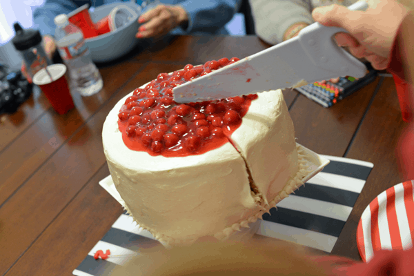Person using a plastic knife to cut a layer cake with cherry pie filling on top.