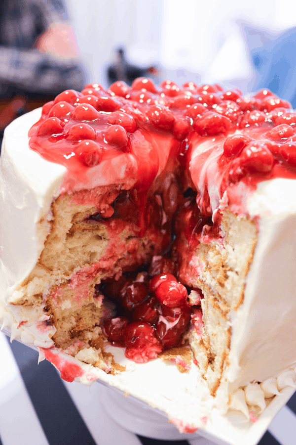 A close up for cherry pie filling oozing out of a vanilla layer cake with cherry pie baked in each layer. 