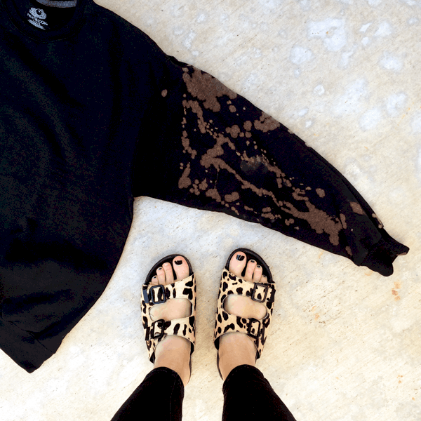 Black sweatshirt laying flat on the ground with one bleach splattered sleeve showing with a woman's feet next to it in sandals.