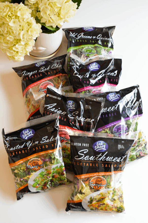 Bags of salad mix on a table next to a vase of white hydrangeas. 