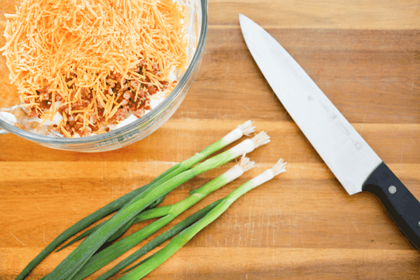 Cutting board with 4 green onions laying next to a knife and a bowl holding ingredients.