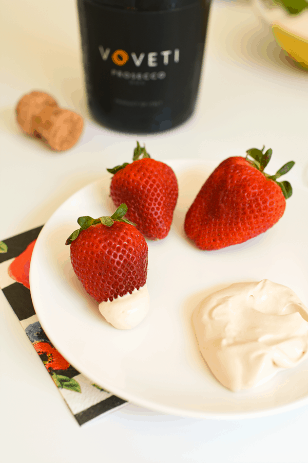 Fresh strawberries and romanoff dip on a white plate in front of a bottle of prosecco.
