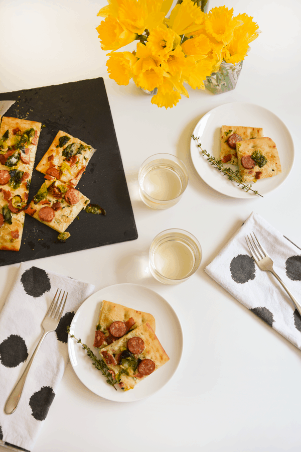 Slices of Goat cheese pizza with sausage, bacon, Brussels sprouts and parmesan cheese on plates and a cutting board surrounded by glasses of wine, napkins and forks. 