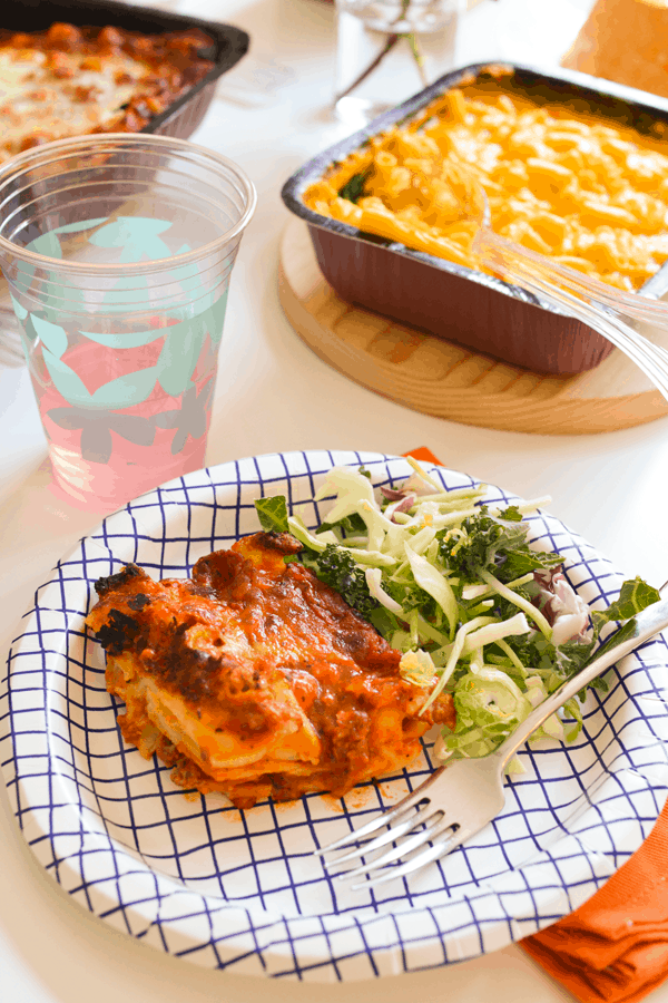 Decorative paper plate with a portion of lasagna and some salad next to a drink and other food on a table. 