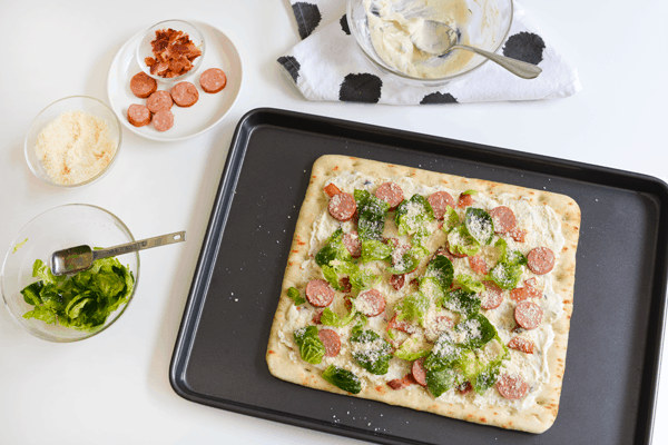 Prepared pizza on a baking sheet next to bowls of ingredients on a white counter top.