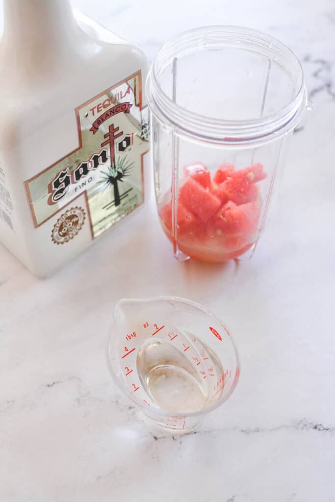 A small measuring cup with tequila next to a blended holding pieces of fresh watermelon.