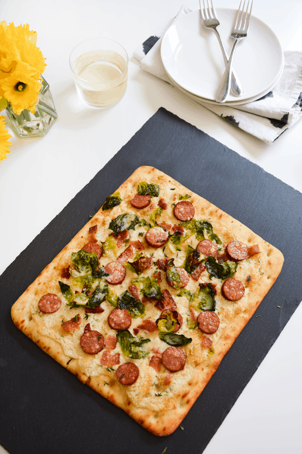 Goat cheese pizza on a black cutting board next to plates and utensils, napkins and a glass of wine.