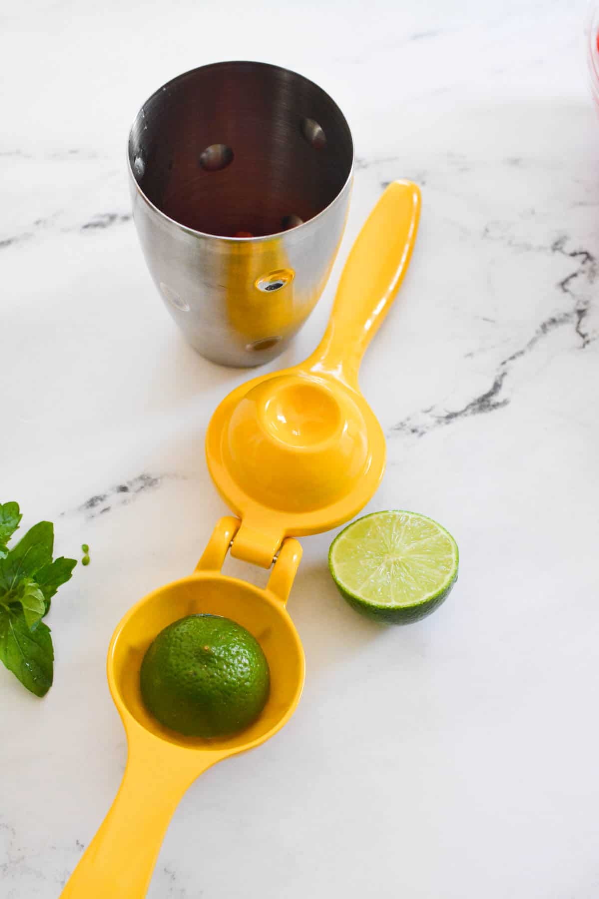Yellow citrus squeezer with half a lime in it on the counter next to a cocktail shaker.
