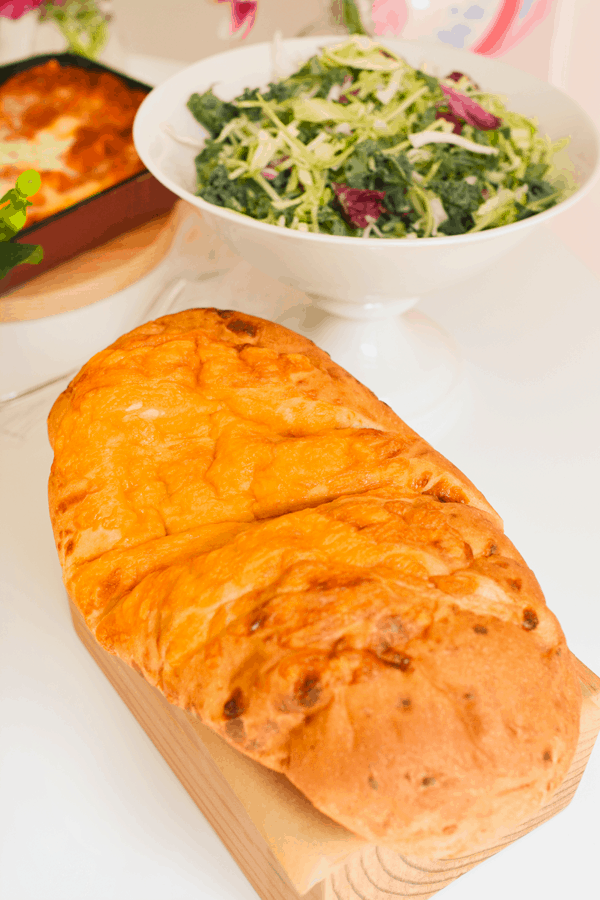 Loaf of cheese bread on a wooden board on a white table next to a salad and other dishes.