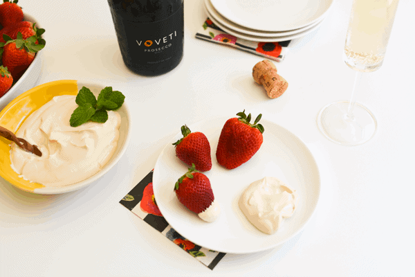 Fresh strawberries and strawberries romanoff dip on a white plate next to a bowl filled with dip and a bottle of prosecco.