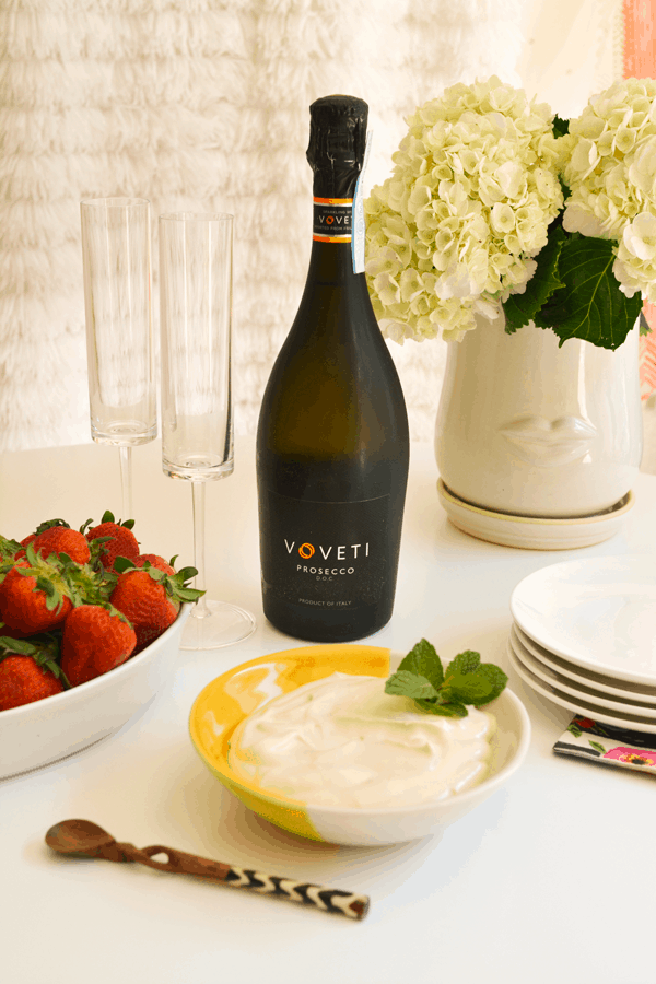A bottle of prosecco on a whote table surrounded by fresh flowers, plates, wine glasses, fresh strawberries and strawberries romanoff dip.