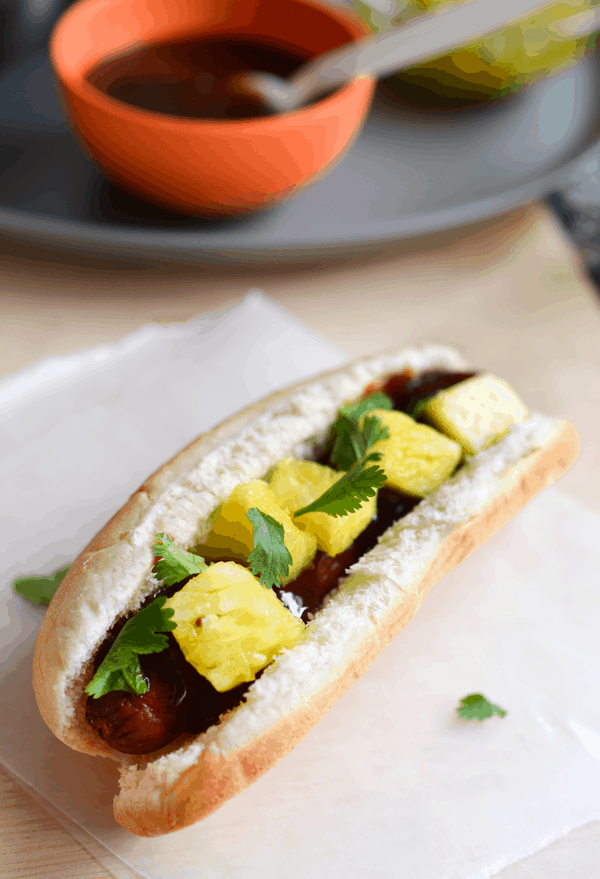A hot dog in a bun topped with bbq sauce, fresh pineapple chunks and cilantro.