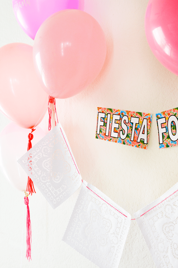 Close up of a banner made from white doilies hanging from pink balloons with tassels and a sign on the wall above it.