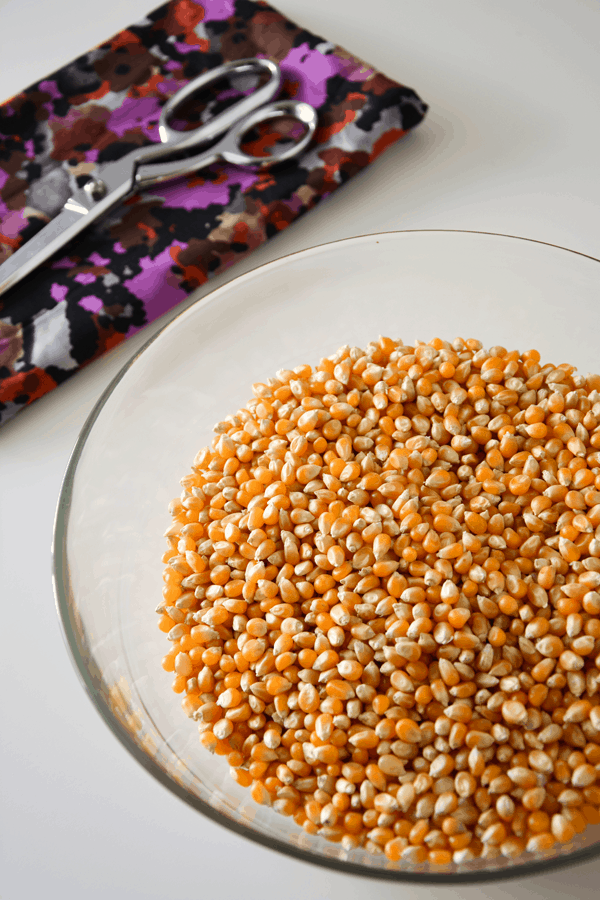 A bowl of popcorn kernels next to a piece of fabric with scissors.
