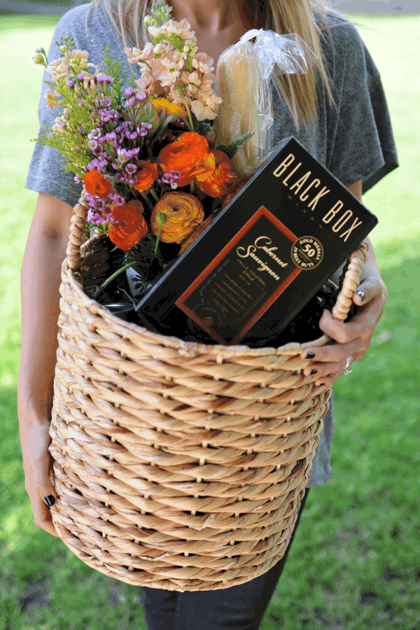 Woman holding a basket of items like fresh flowers and a box of wine for an outdoor party. 