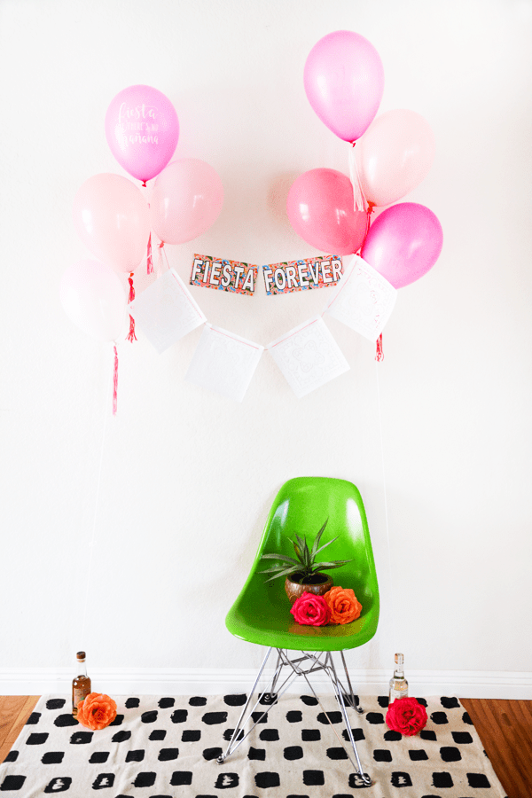 Pink balloons holding a white papel picado style banner and a sign on the wall that says "fiesta forever" floating above a bright green chair. 