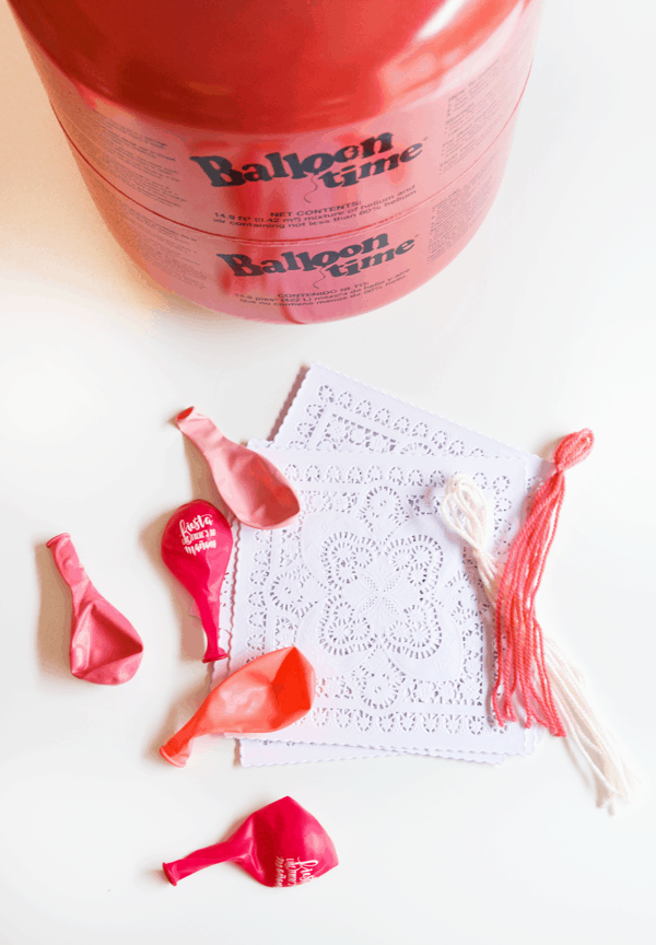 A helium tank to blow up balloons on a table next to yarn tassels, pink balloons and white doilies.