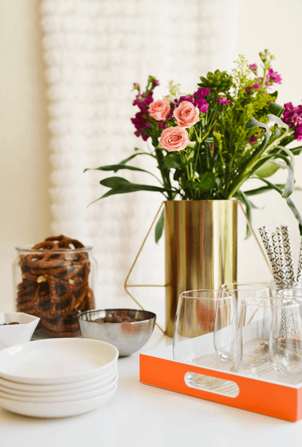 A gold vase with flowers on a table next to a tray of wine glasses and other party supplies. 