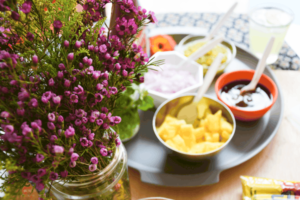 Small purple flowers on a food table with hot dog toppings in the background.