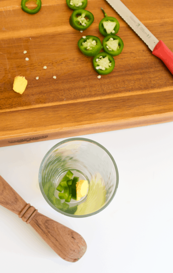 A glass with pieces of sliced jalapeno and ginger in it next to a cutting board with a knife. 